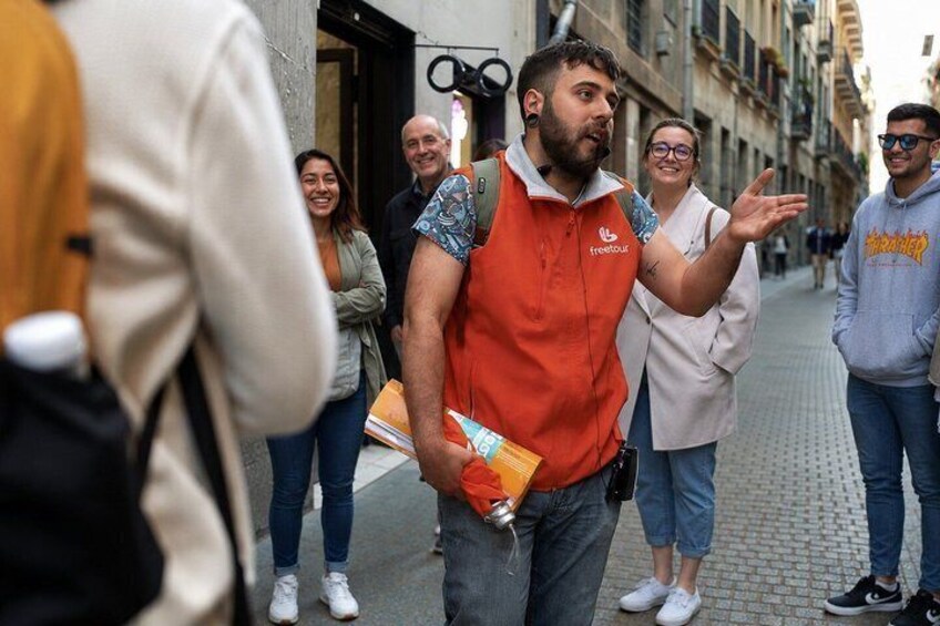 A person in an orange vest gesturing, with a crowd of people and buildings in the background. 1