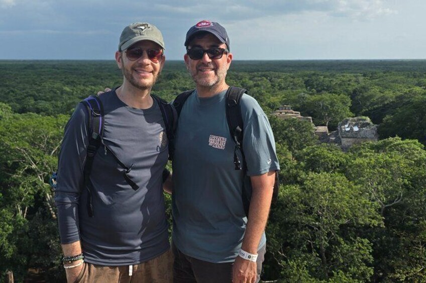 Two people standing on a hill with a forested landscape and ancient ruins in the background. 5