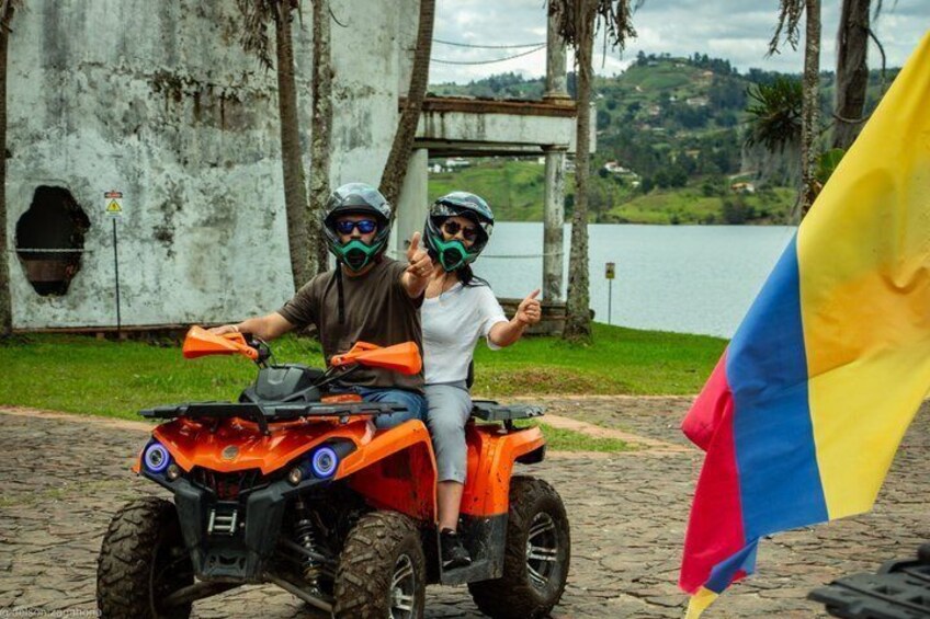 Two people on an orange ATV, a flag, and a body of water in the background. 1
