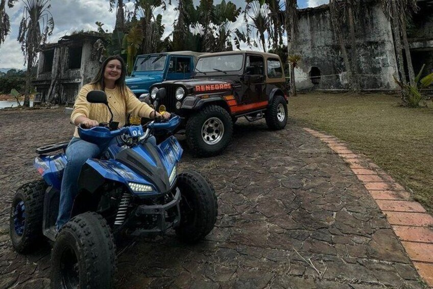 A person on a blue quad bike, a black and orange off-road vehicle, and a stone-paved area with trees and a building in the background. 4