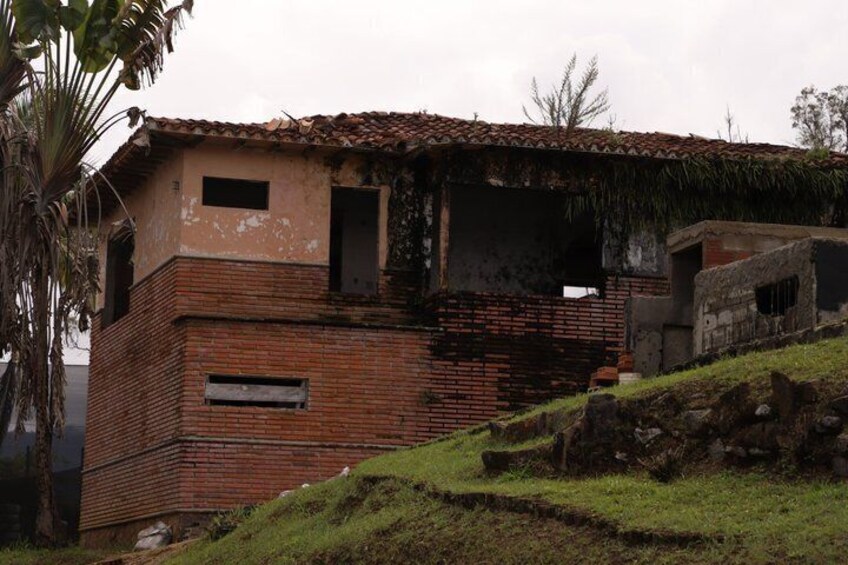 A dilapidated brick building with a tiled roof, surrounded by grass and a palm tree. 2