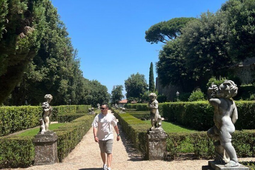 A person walking on a gravel path in a garden with statues and neatly trimmed hedges. 4