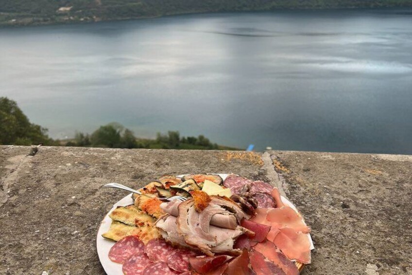 A plate of cured meats and cheeses on a concrete surface with a lake in the background. 2