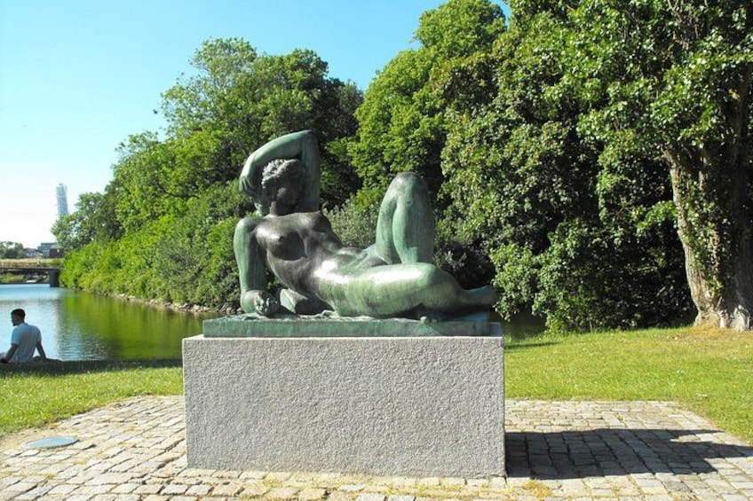A bronze sculpture of a reclining figure in a park with a river and trees in the background. 4