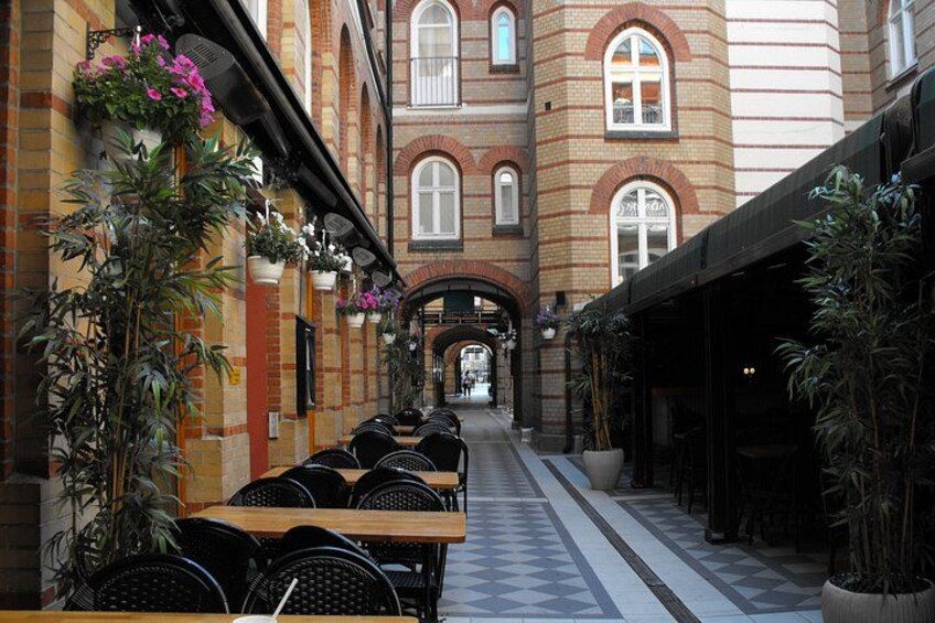 A covered outdoor seating area with tables and chairs, potted plants, and a brick building with arched windows. 5