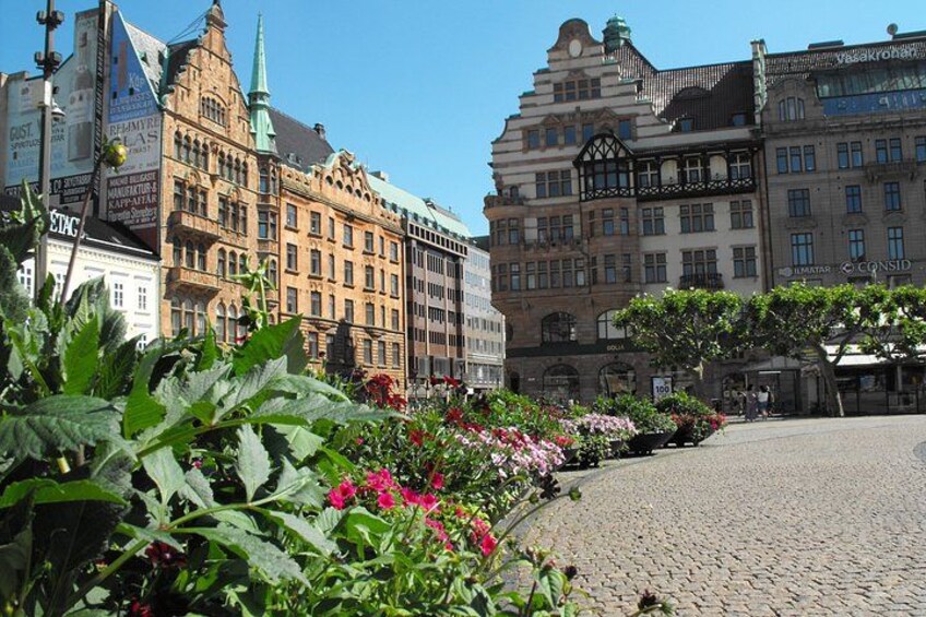 A cobblestone square with historic buildings, a church spire, and flowering plants. 2