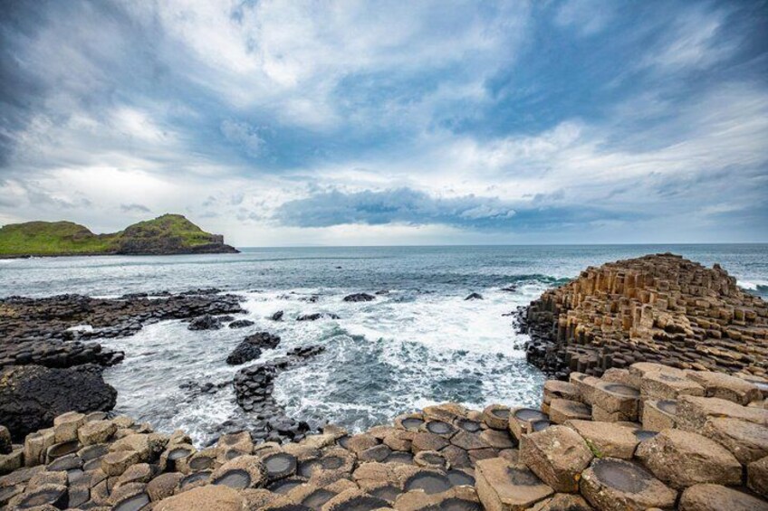 A rocky coastline with waves crashing against the shore, a distant green hill, and a stone structure on the right. 4