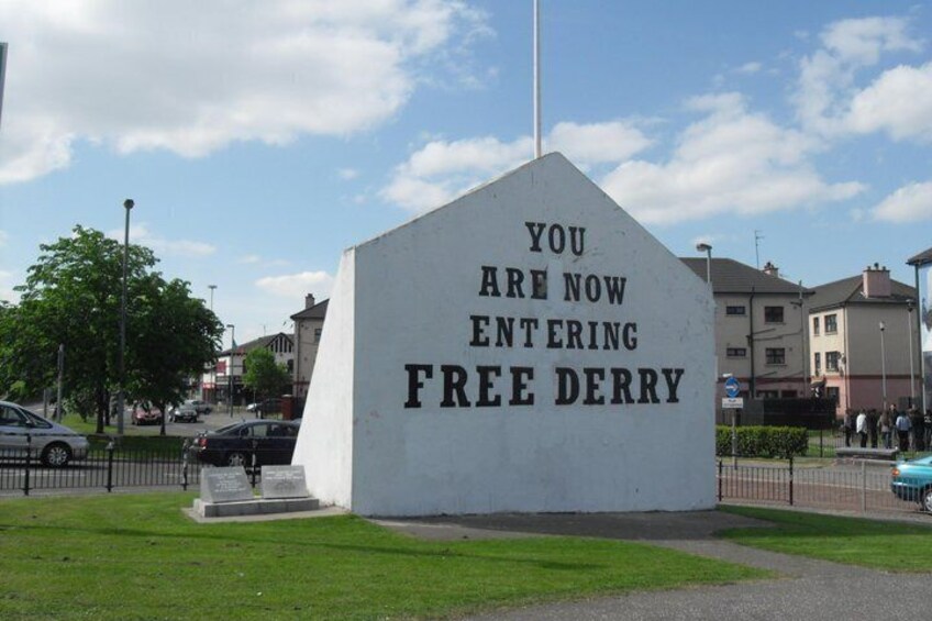 A white monument with the inscription 'YOU ARE NOW ENTERING FREE DERRY' on a grassy area with buildings in the background. 3