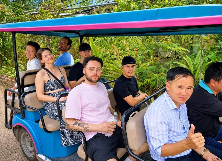 A group of people seated in a colorful trolley with a blue and pink canopy, surrounded by lush greenery. 4