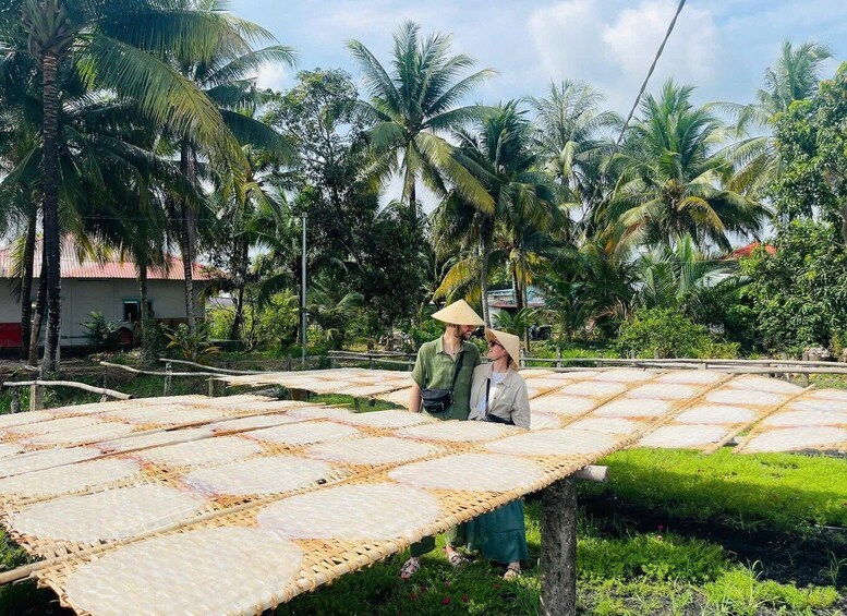 Two people standing on a woven mat in a tropical setting with palm trees and a house in the background. 2