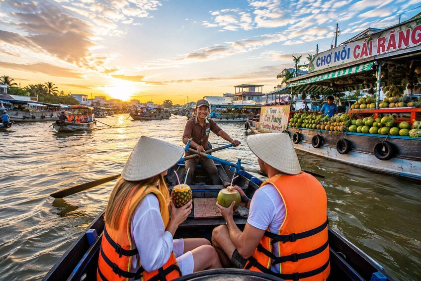A boat with two passengers wearing life jackets and conical hats, navigating a river with a floating market on the right. 1