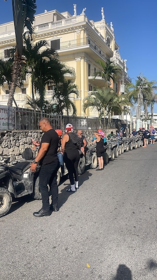A group of people in black attire standing in front of ATVs, with a building and palm trees in the background. 2