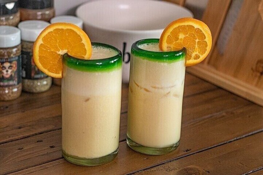 Two glasses of a creamy beverage with orange slices on the rim, placed on a wooden surface with spices in the background. 4