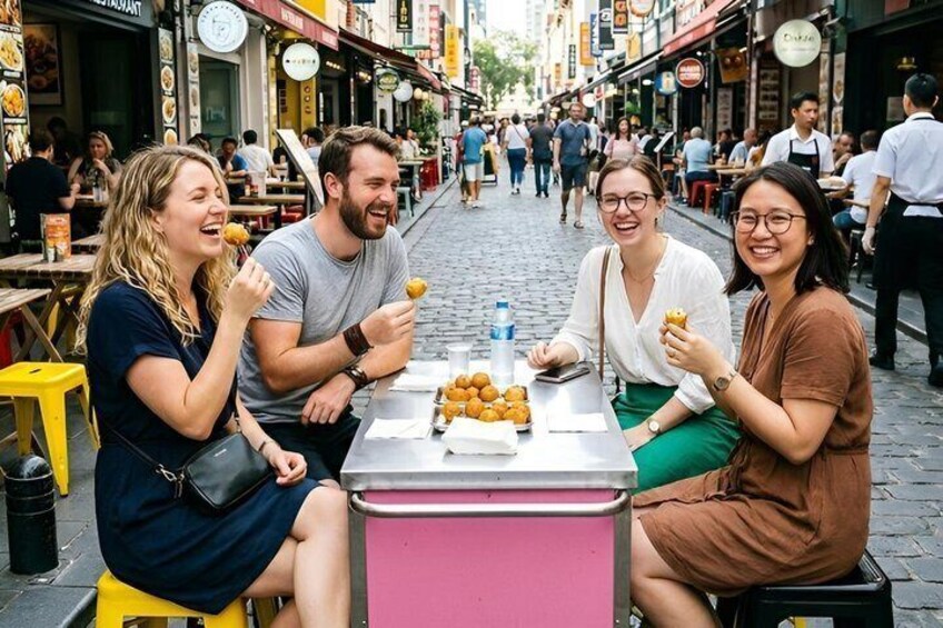 A group of people sitting at a table with food, in a busy outdoor market area. 1