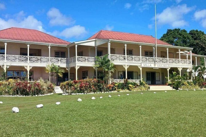 A two-story building with a red roof and white balcony railings, surrounded by a well-maintained lawn and tropical plants. 3