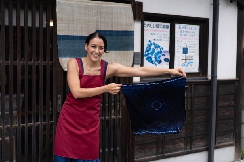 A person holding a blue fabric with a circular pattern, standing in front of a building with wooden slats and a window displaying posters. 5