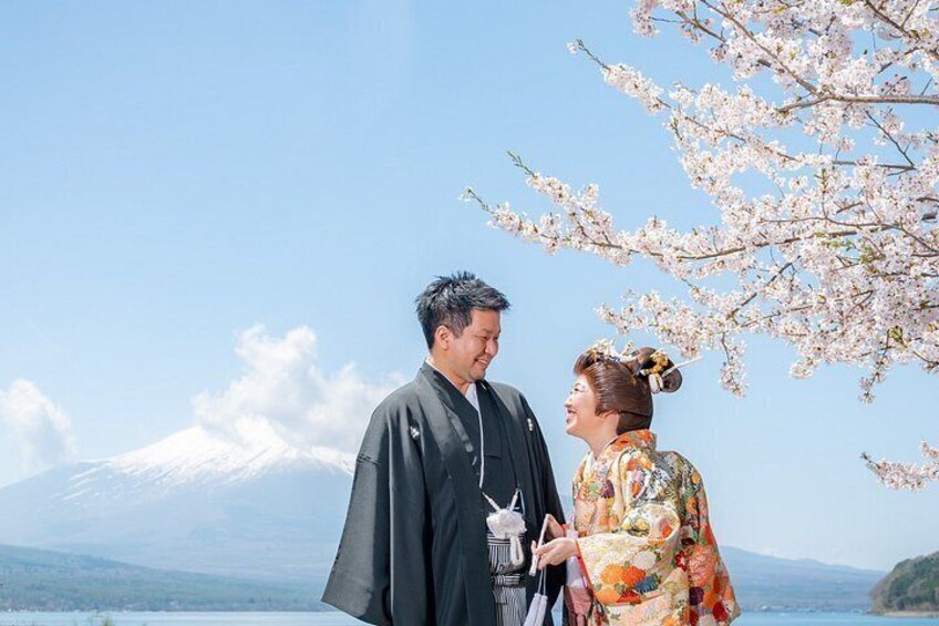 A couple in traditional attire standing by a cherry blossom tree with a mountain in the background. 2