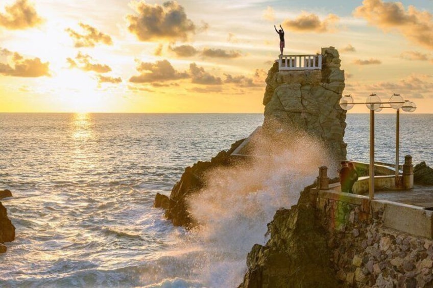 A rocky coastline with waves crashing against it, a person standing on a ledge, and a sunset in the background. 5