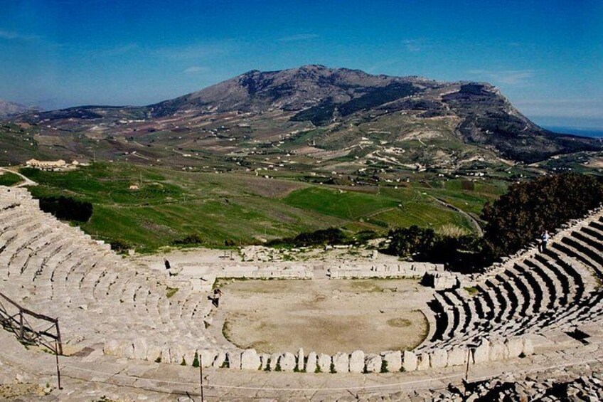 An ancient amphitheater with tiered seating and a central stage, surrounded by a mountainous landscape. 4