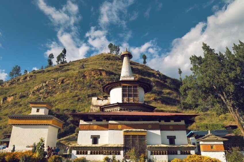 A white stupa with a conical roof, surrounded by traditional buildings with red and white walls, set against a hillside with sparse vegetation. 2