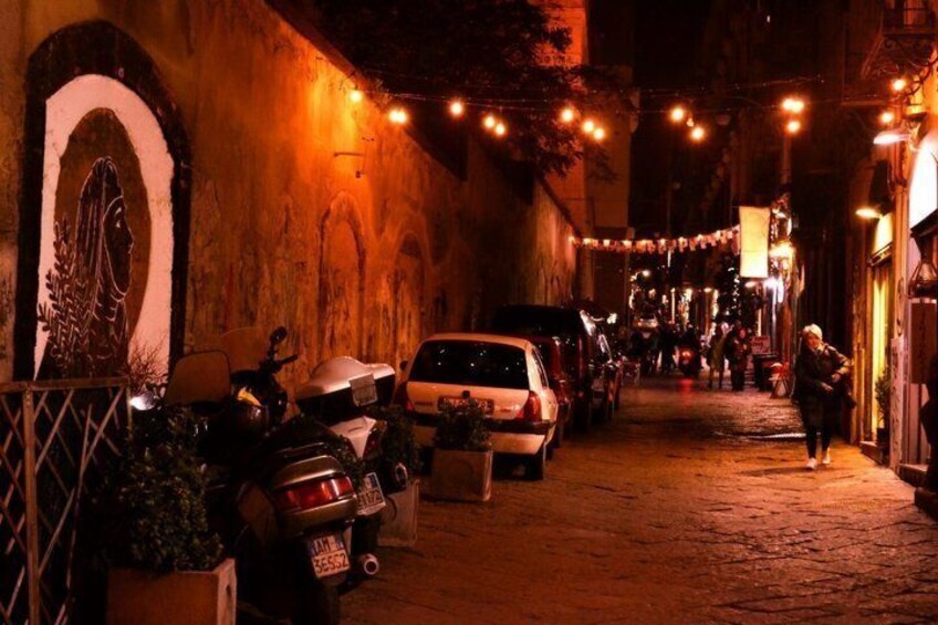 A narrow street at night with parked cars, a motorcycle, and a person walking. 2