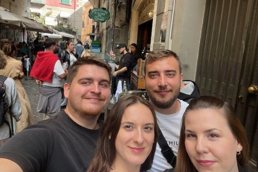 A group of people taking a selfie in a narrow street with shops and pedestrians in the background. 4