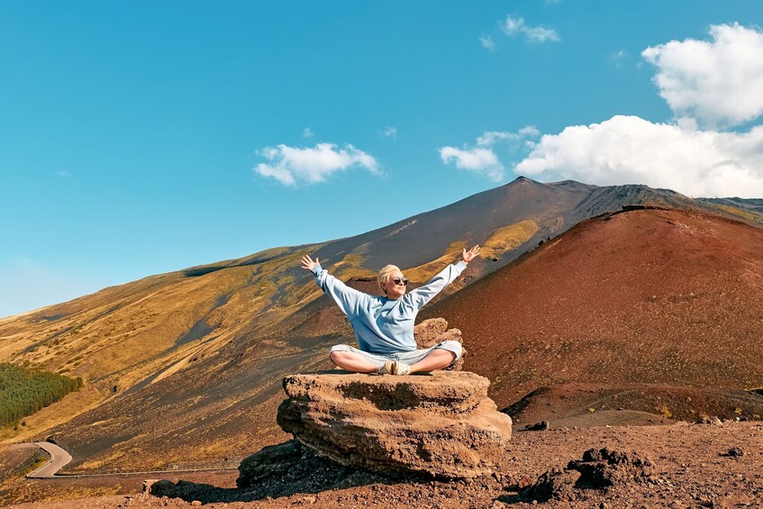 A person sitting on a rock with arms raised, a mountain in the background, and a clear sky. 2