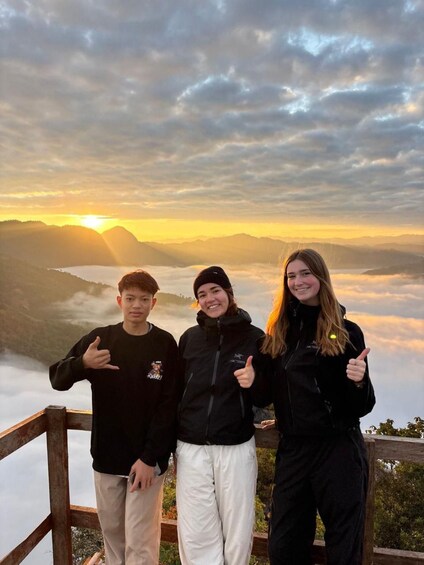 Three people giving thumbs up on a wooden platform with a mountainous landscape and a sunrise in the background. 2