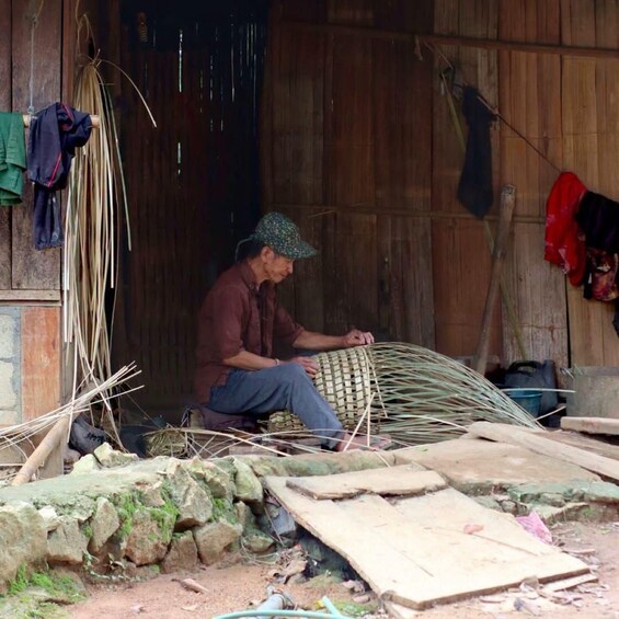 A person sitting on a woven mat, working with bamboo sticks in a rustic setting with wooden walls and scattered construction materials. 4