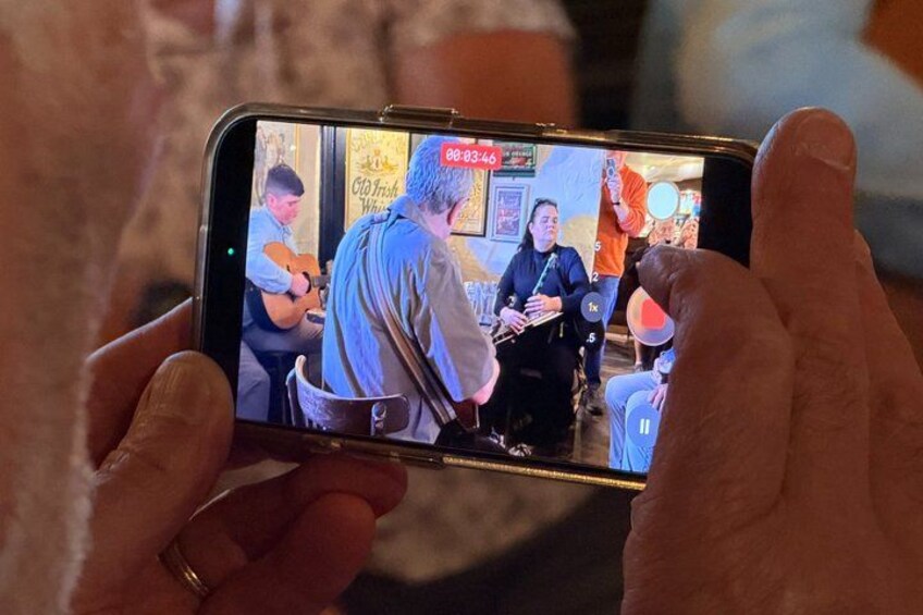 A person holding a smartphone, recording a scene with a guitar player and a seated individual. 5