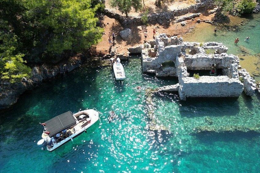 Eine Luftaufnahme einer Küstenlandschaft mit klarem türkisblauem Wasser, einem Boot, das an Ruinen liegt, und Menschen, die im Wasser schwimmen. 1