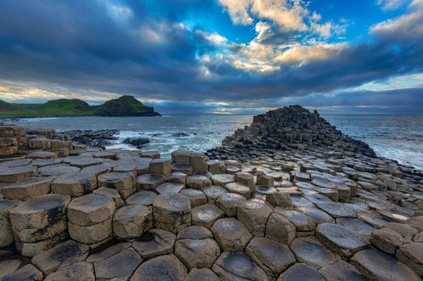 A hexagonal stone structure by the sea, a rocky coastline, and distant hills. 1
