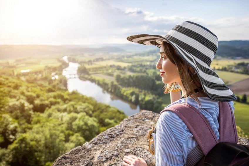 A person wearing a striped hat and a backpack, overlooking a river and green landscape. 1