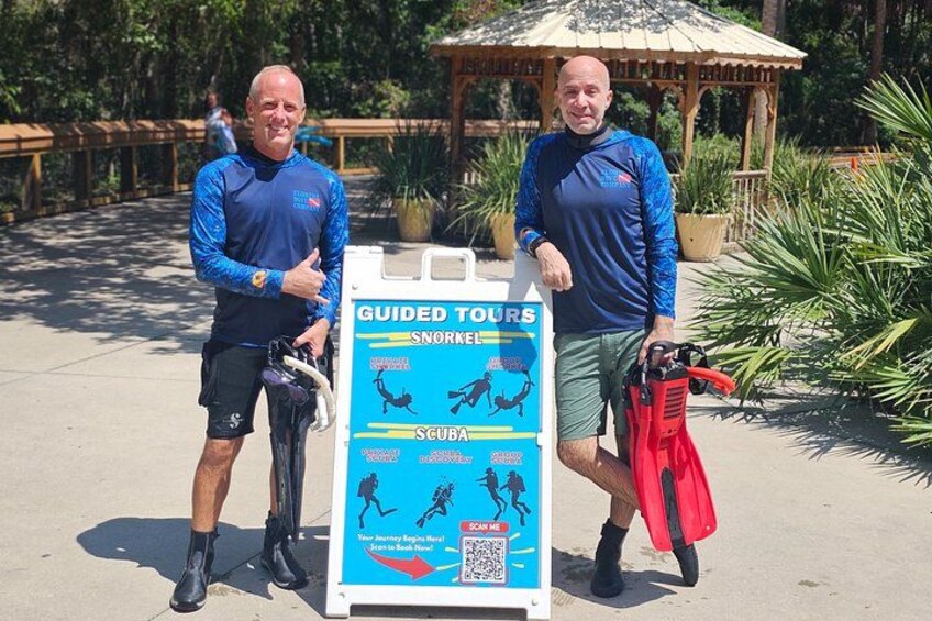 Two men standing by a sign for guided tours, one holding a snorkel and the other a scuba diving mask. 5