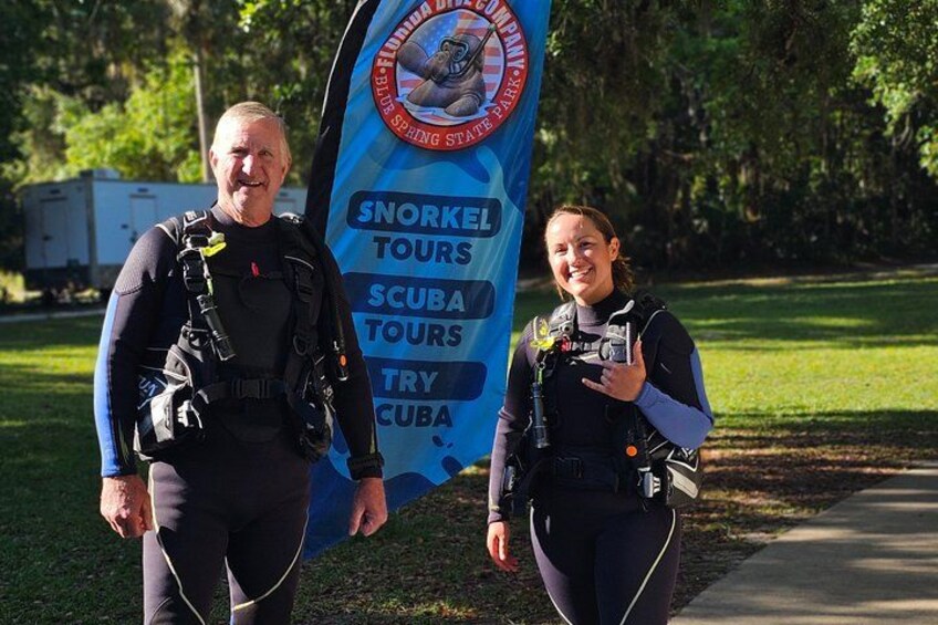 Two people in scuba diving gear standing in front of a banner advertising snorkeling, scuba, and try-cuba tours. 4