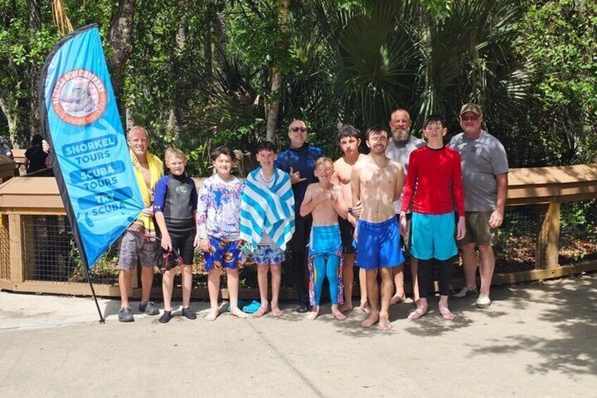 A group of people standing on a concrete surface with a blue flag featuring a logo and text, surrounded by greenery. 2