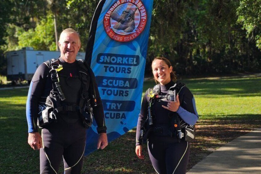 Two people in scuba diving gear standing in front of a banner advertising snorkeling, scuba, and try-cuba tours. 5