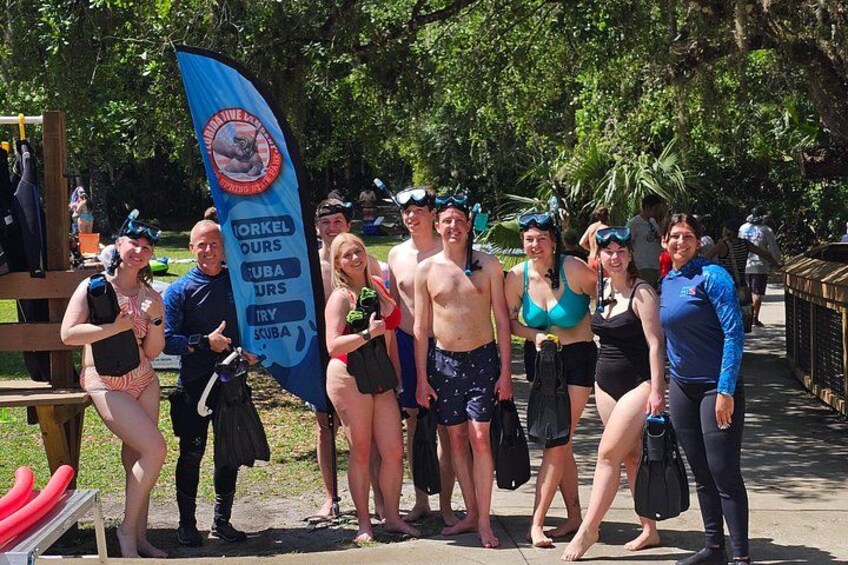A group of people in swimwear and snorkeling gear posing for a photo with a flag that reads 'CORKEL TOURS UBA JURS'. 1