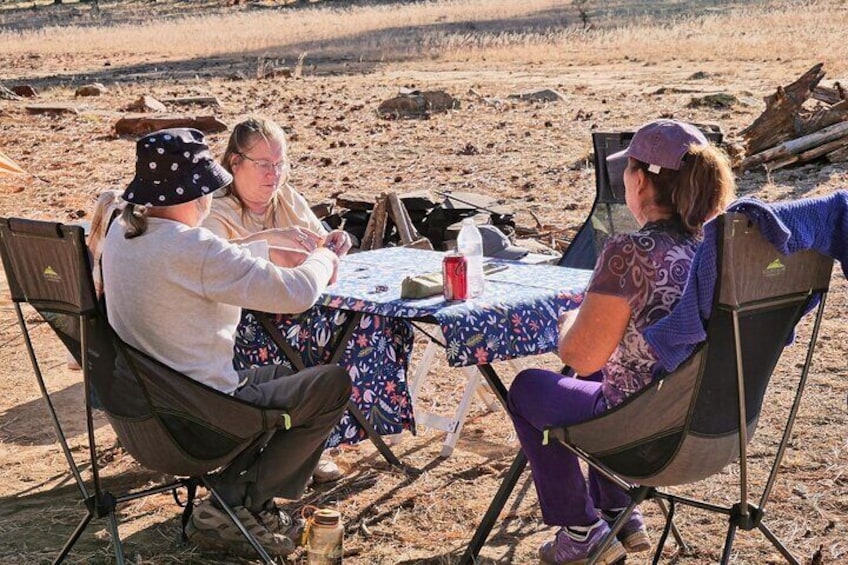 Three people sitting at a table outdoors with camping chairs, a tablecloth, and a bottle. 1