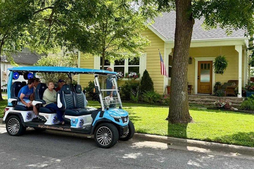 A golf cart with passengers parked in front of a yellow house with a flag. 1