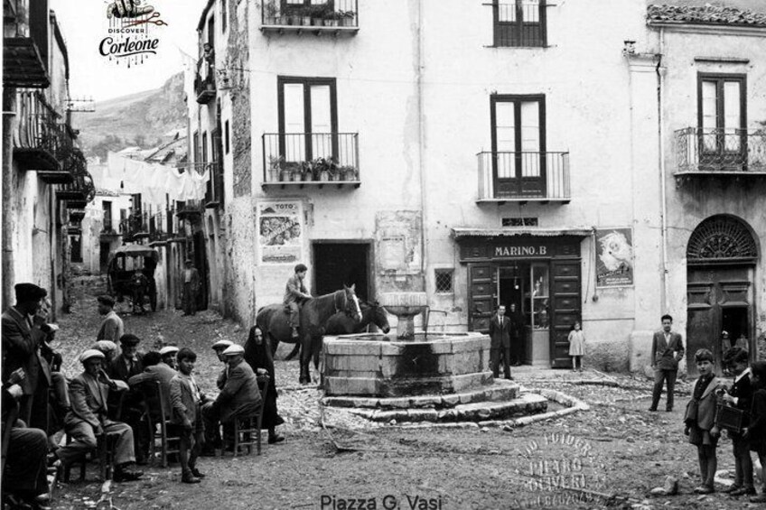 A historical black and white street scene with people, a horse, and buildings. 1