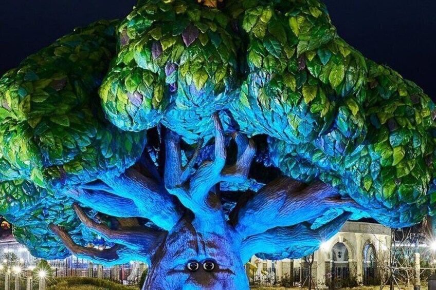 A large tree with blue lighting, surrounded by a park with a building in the background. 4