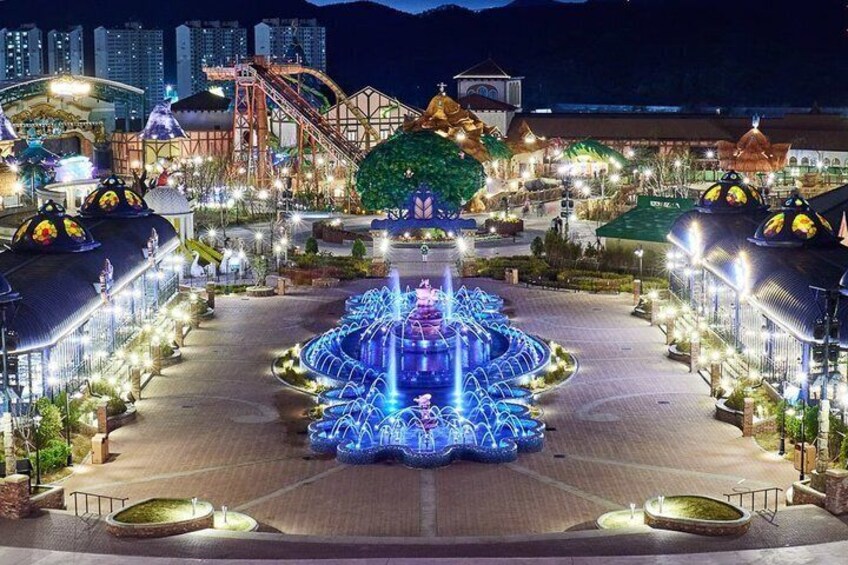 A brightly lit amusement park at night with a central fountain, roller coaster, and traditional buildings. 2
