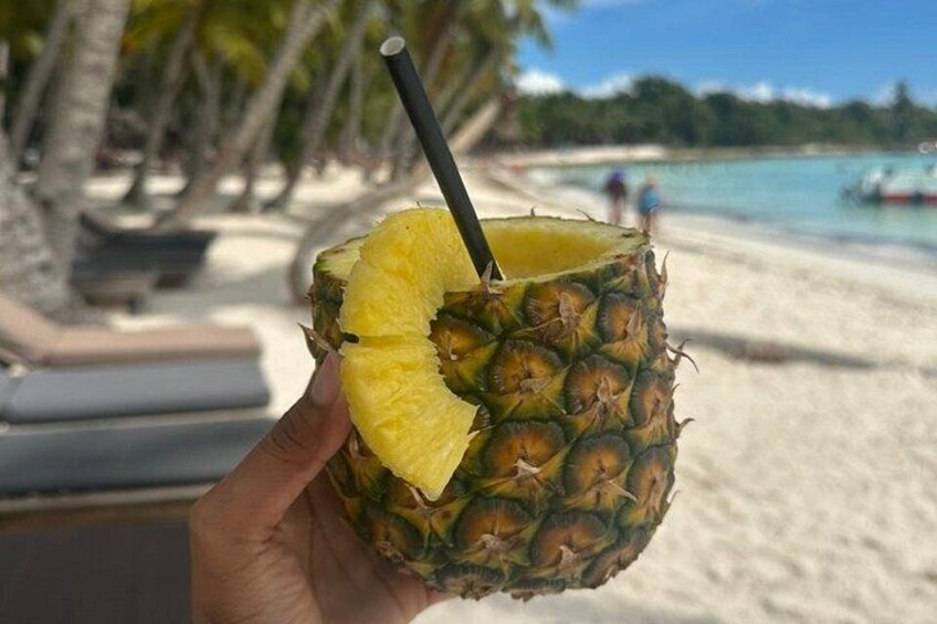 A hand holding a pineapple with a straw, set against a beach with palm trees and a boat in the background. 2