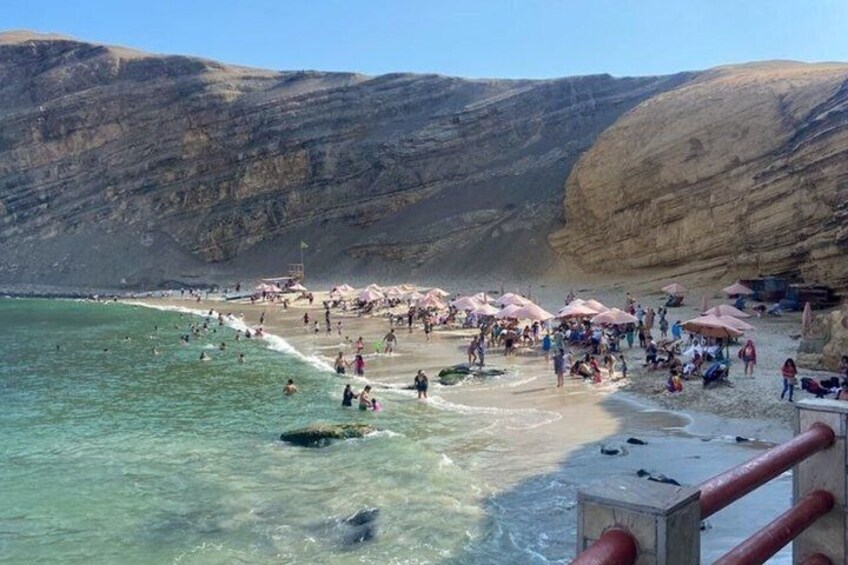 A beach with people swimming and sunbathing, umbrellas, and rocky cliffs in the background. 4