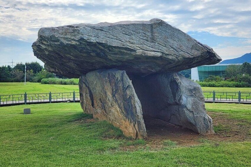 A large standing stone in a grassy field with a modern building in the background. 1