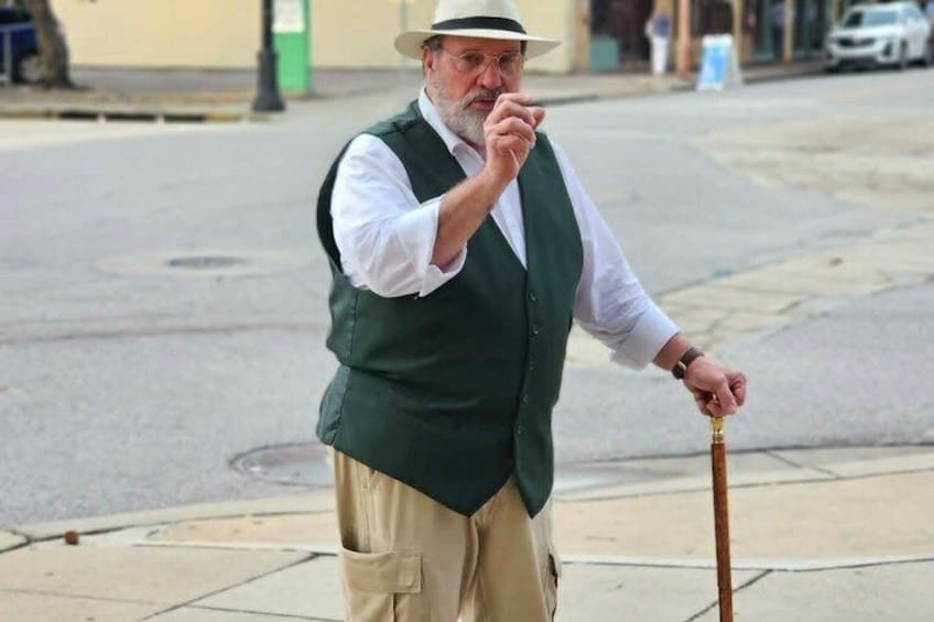 An elderly man in a green vest and beige pants, holding a cane, standing on a sidewalk. 1