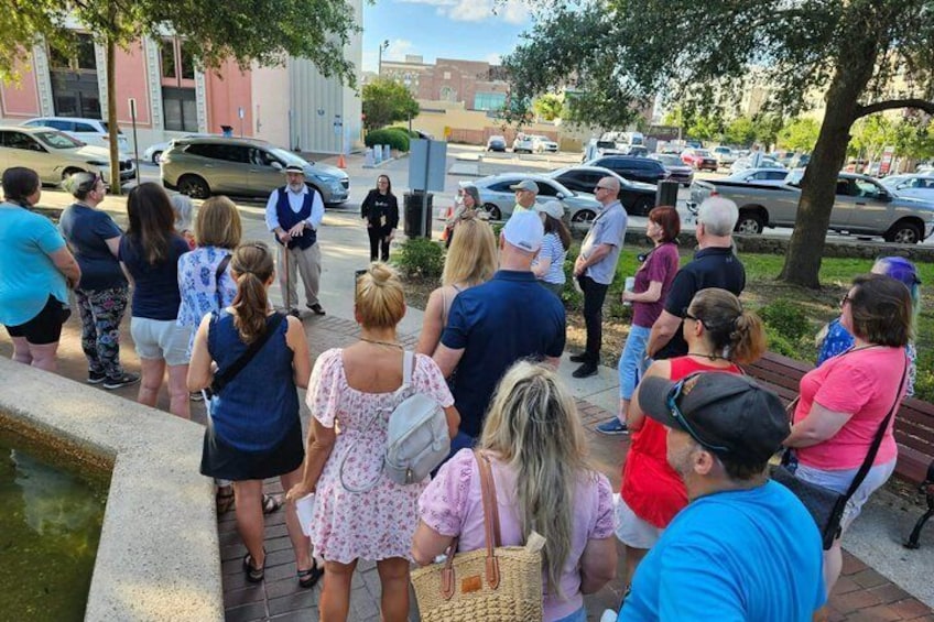 A group of people standing on a sidewalk, some with bags, near a water feature and parked cars. 5