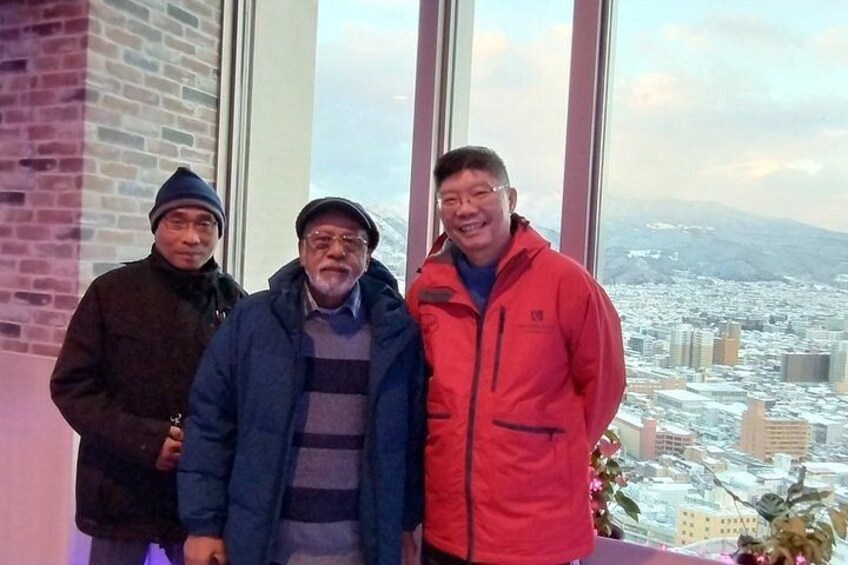 Three people standing indoors with a cityscape view through large windows. 3