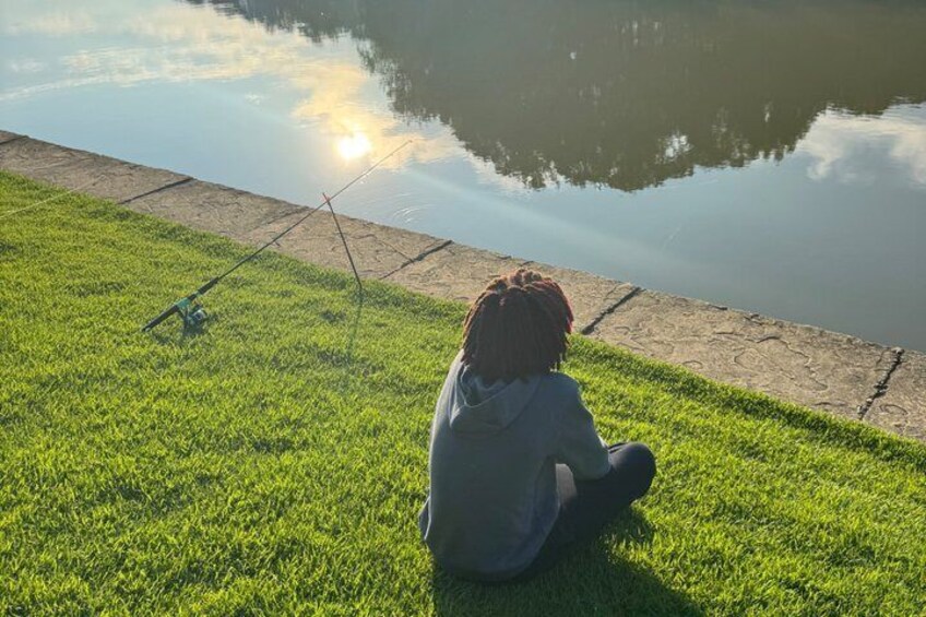 A person sitting on grass by a calm body of water with fishing rods. 5